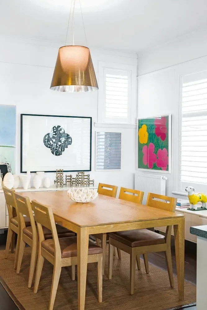 Bright dining room with wooden table and chairs, white walls, and colorful wall art under a copper pendant light