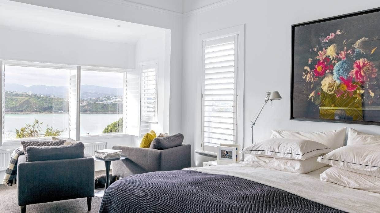 Bright white bedroom with gray chairs, a black bedspread, floral wall art, and windows overlooking water.