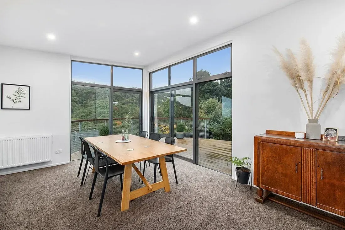 Modern dining room with wooden table, black chairs, large corner windows, and a sideboard with decor.