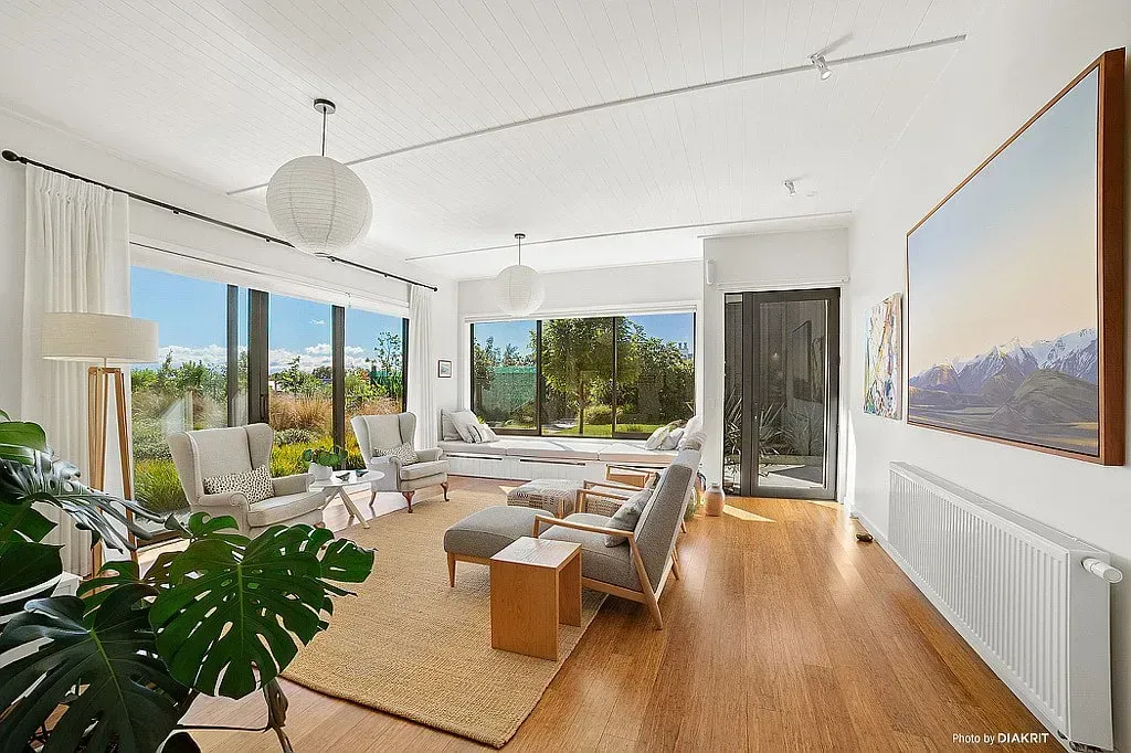 Bright modern living room with large windows, white furniture, wood floors, and a large plant and artwork.
