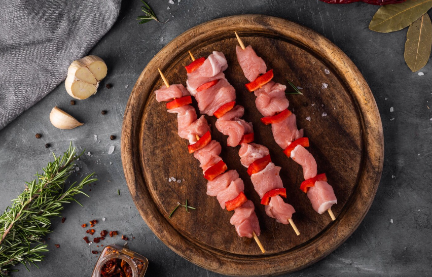 Spiedini di carne cruda con peperone rosso su un piatto di legno, contornato da aglio e rosmarino su una superficie scura.