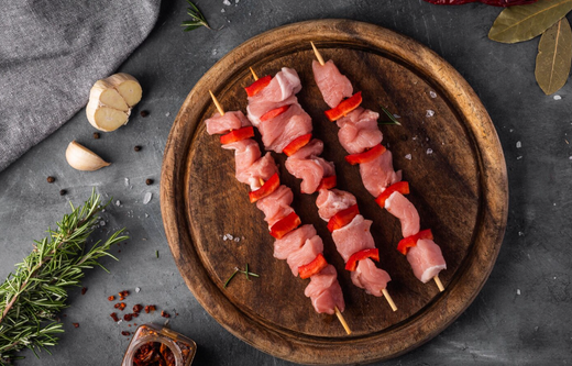 Spiedini di carne cruda con peperone rosso su un piatto di legno, contornato da aglio e rosmarino su una superficie scura.