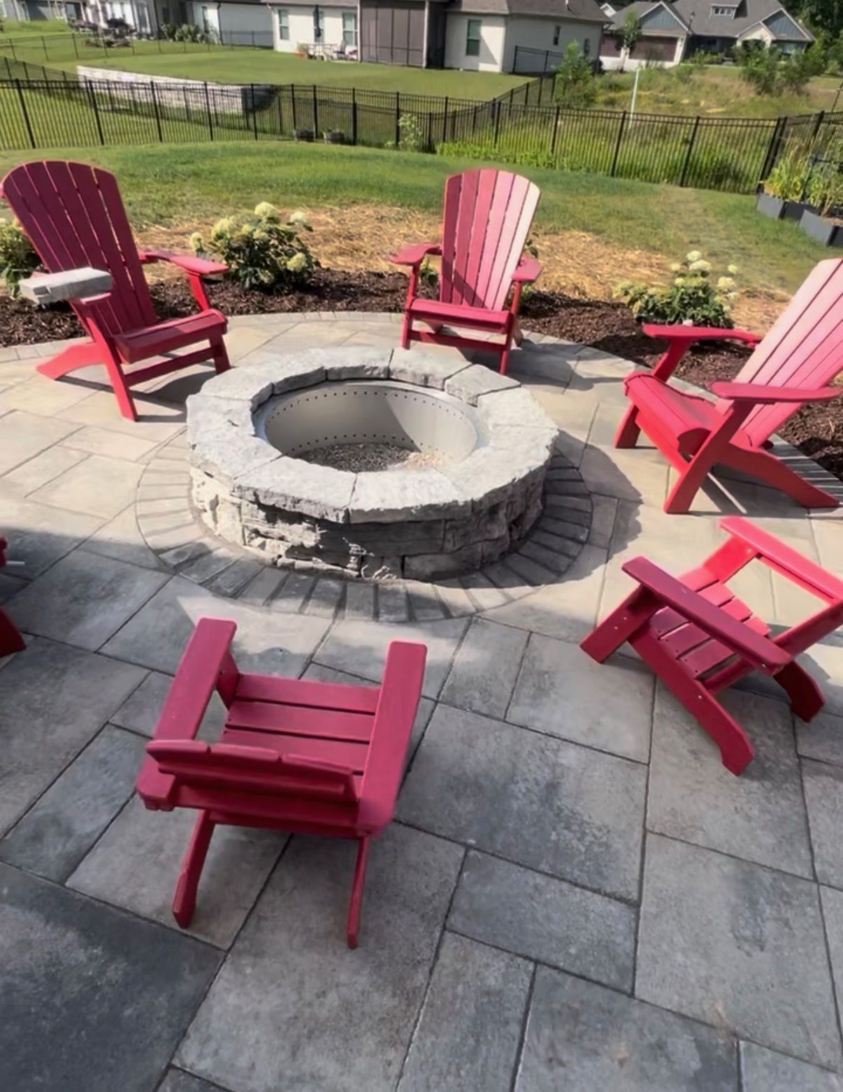Backyard patio with a circular stone fire pit surrounded by five red Adirondack chairs