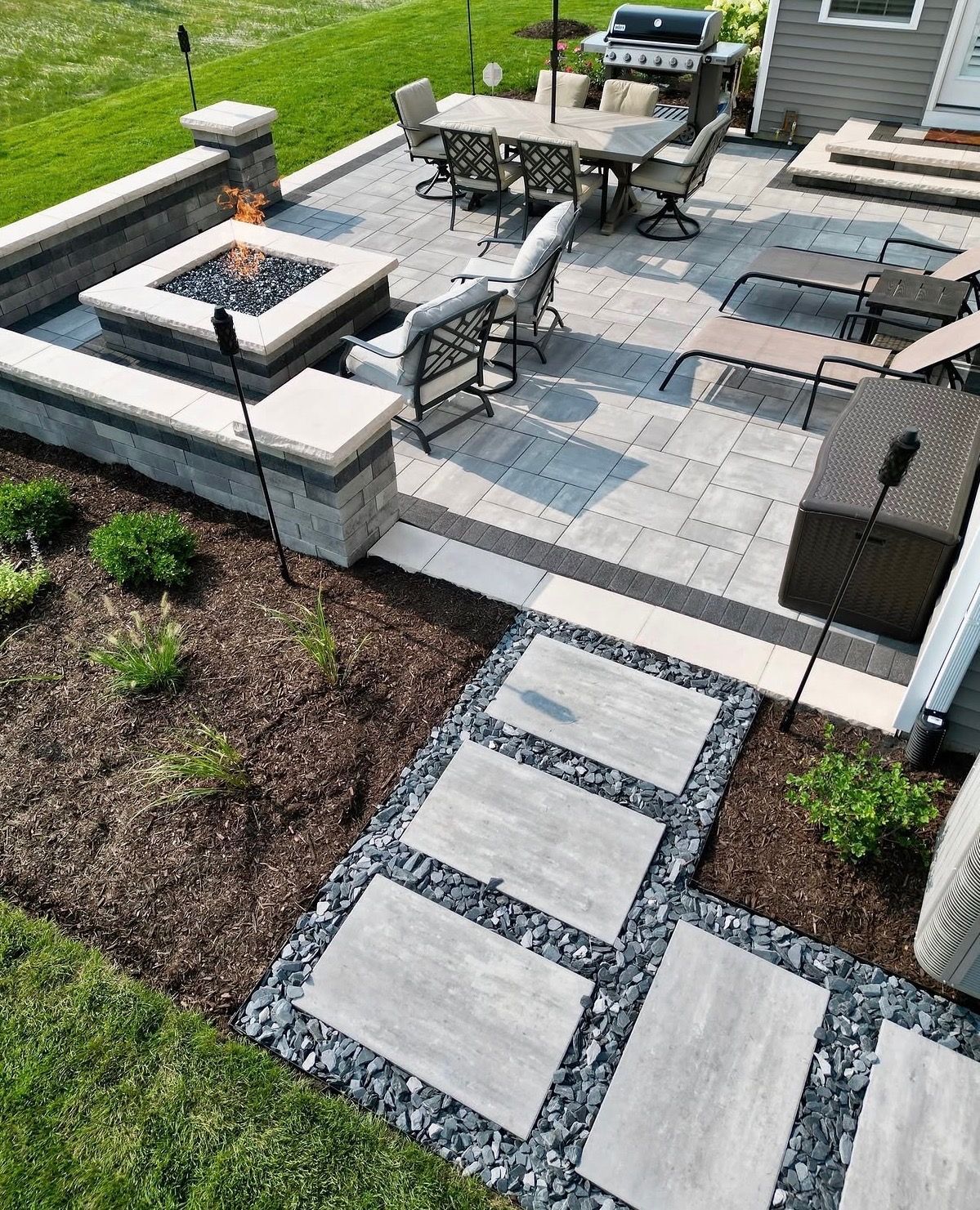 Stone patio with fire pit, dining table, chairs, and a walkway through mulch in a landscaped backyard.