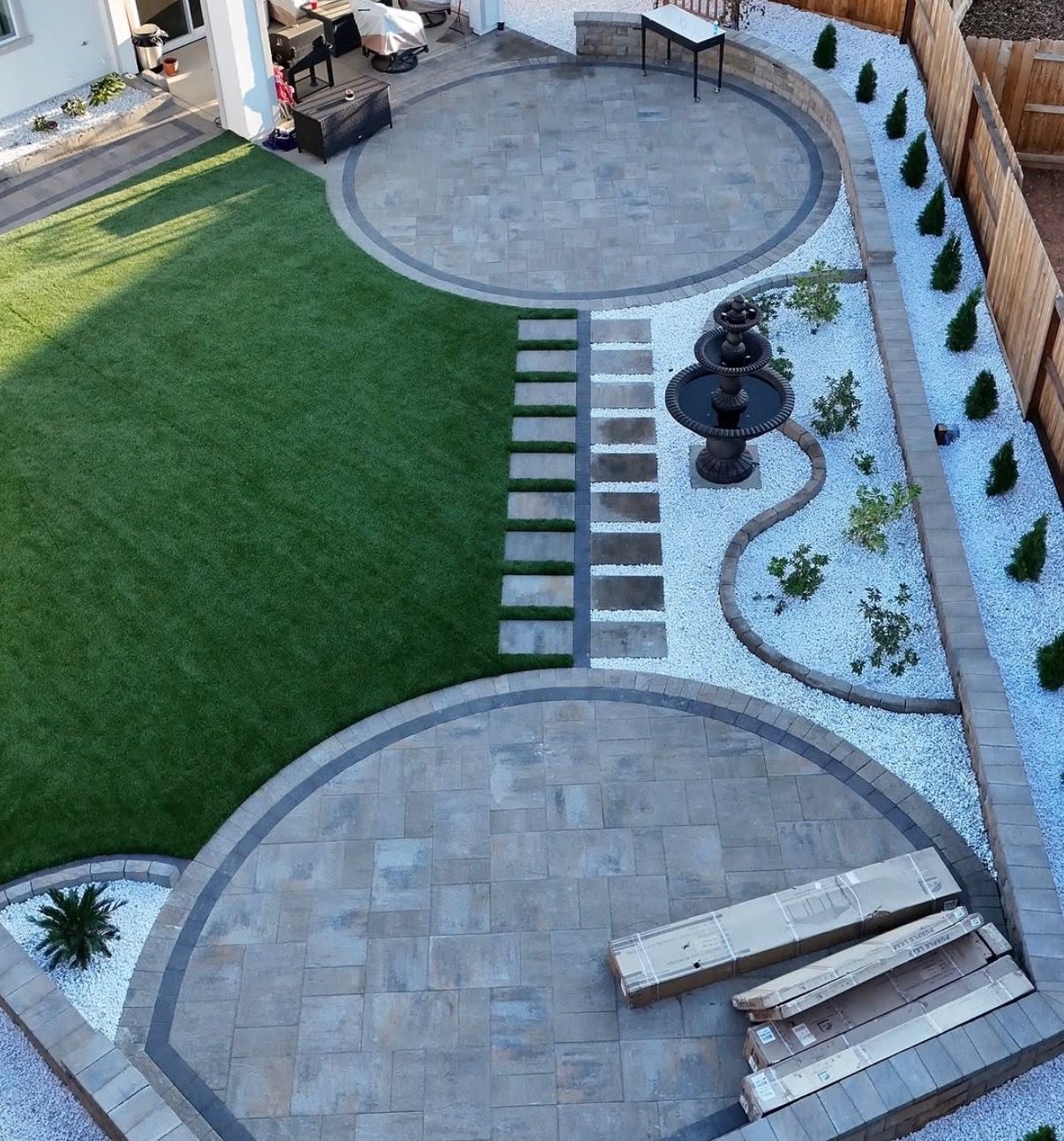 Backyard with curved stone patios, green lawn, stepping stones, fire pit, and pebble garden border.