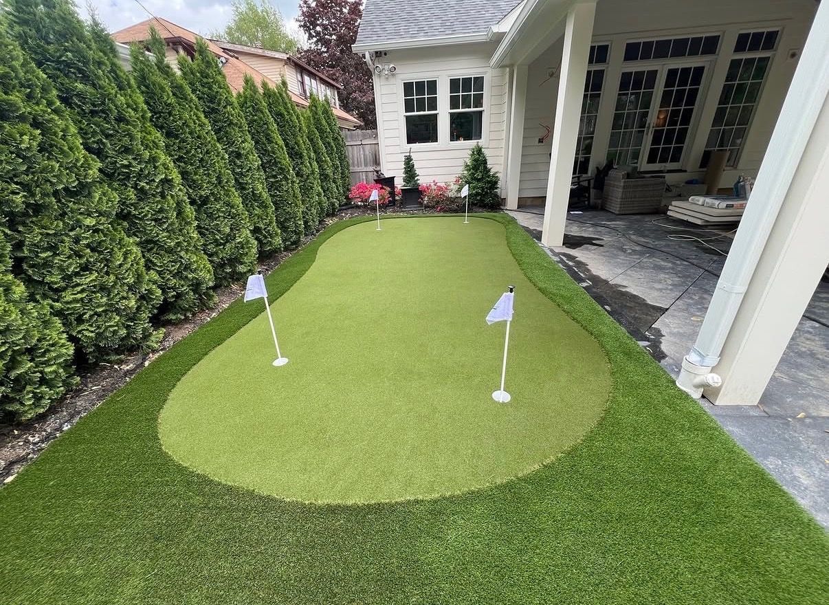 Small backyard putting green with two golf flags beside a house and hedge-lined fence