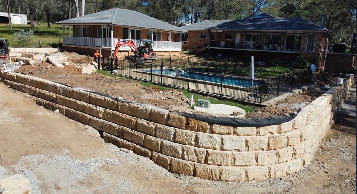 Retaining wall and backyard pool under construction beside a house, with stacked stone and dirt foreground