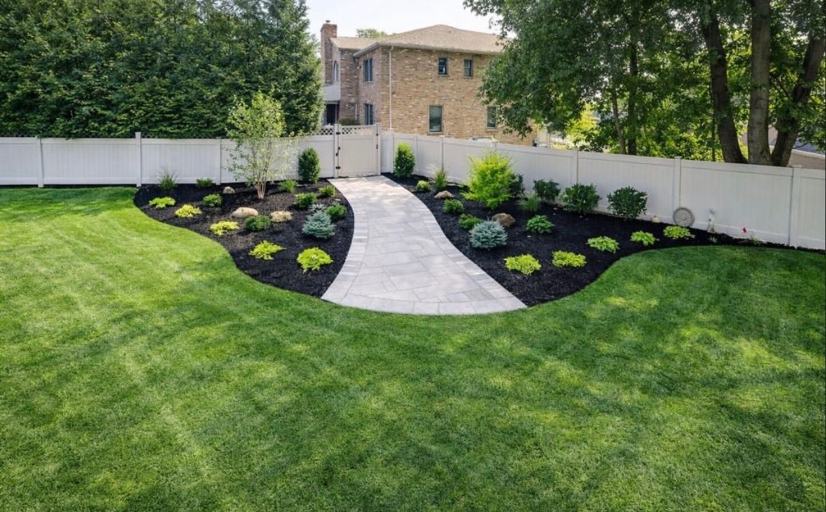 Landscaped backyard with curved stone path, black mulch beds, and a white fence in front of a house