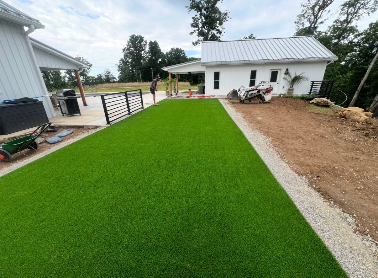 Newly installed bright green artificial turf beside a white house and gravel driveway.
