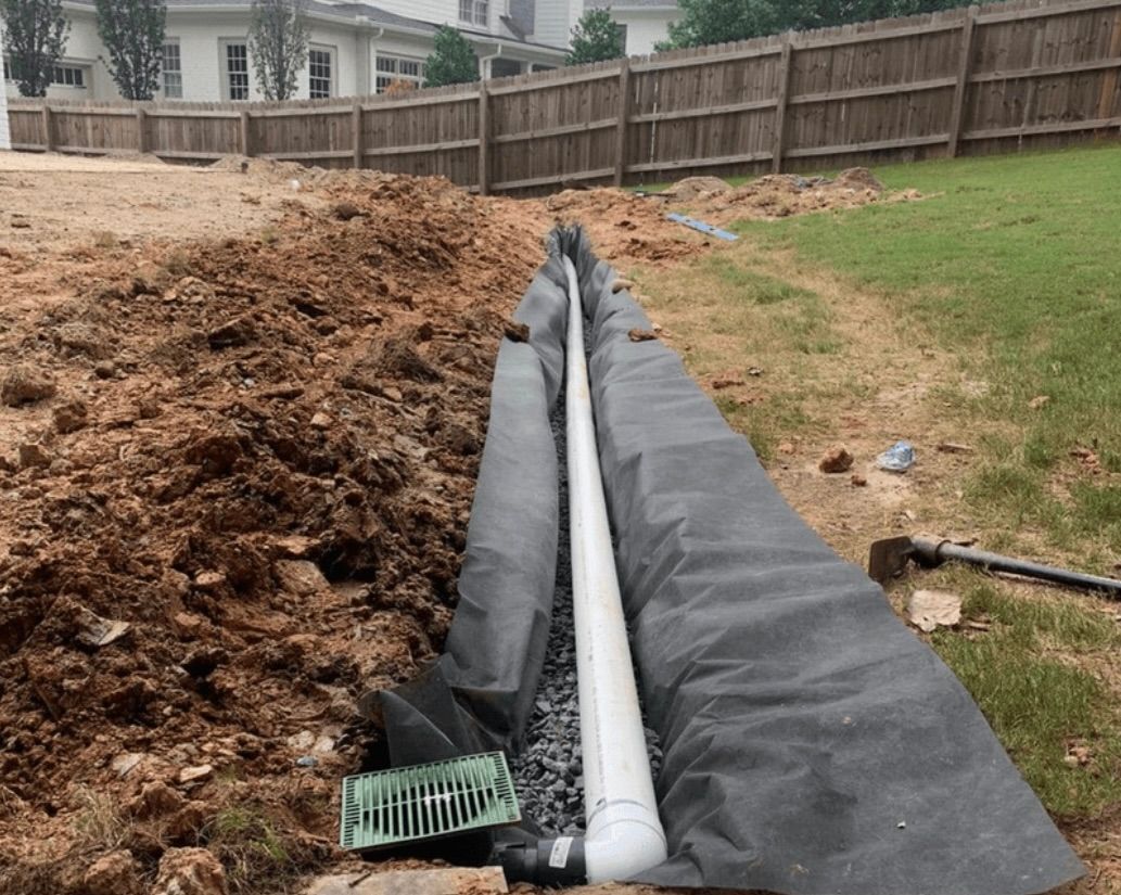 Storm drain pipe lined with black fabric beside a dirt trench in a backyard construction area