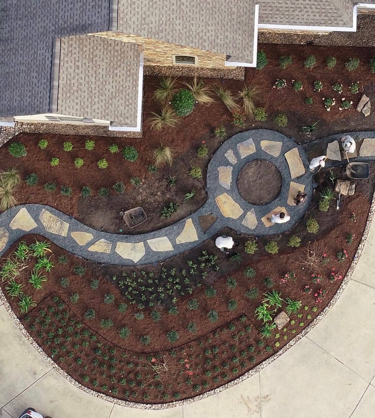 Curved stone path through a mulched garden bed beside a building, with small green plants and a circular patio.