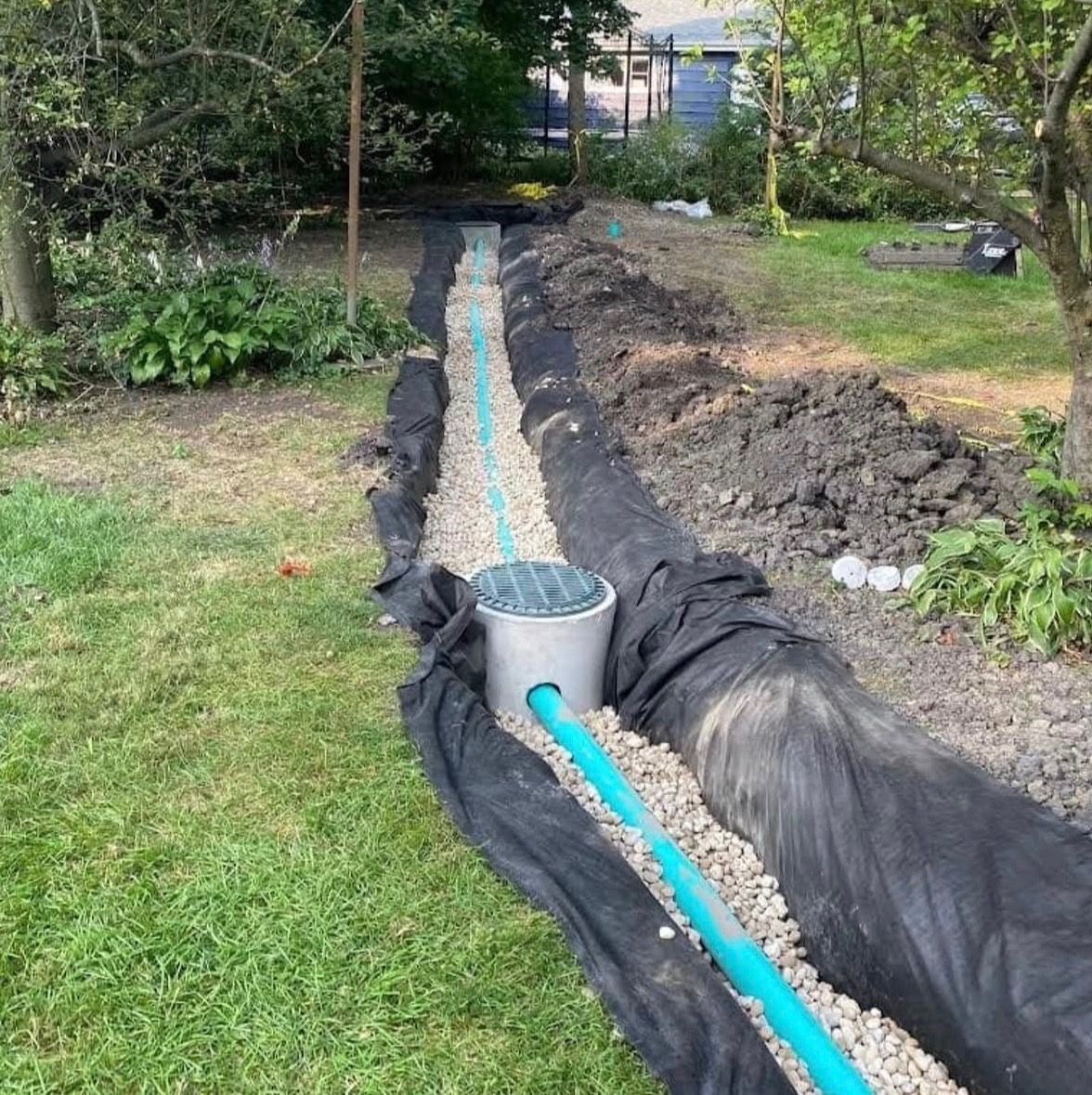 A narrow trench lined with black fabric, a blue pipe, and a bucket in a grassy backyard garden.