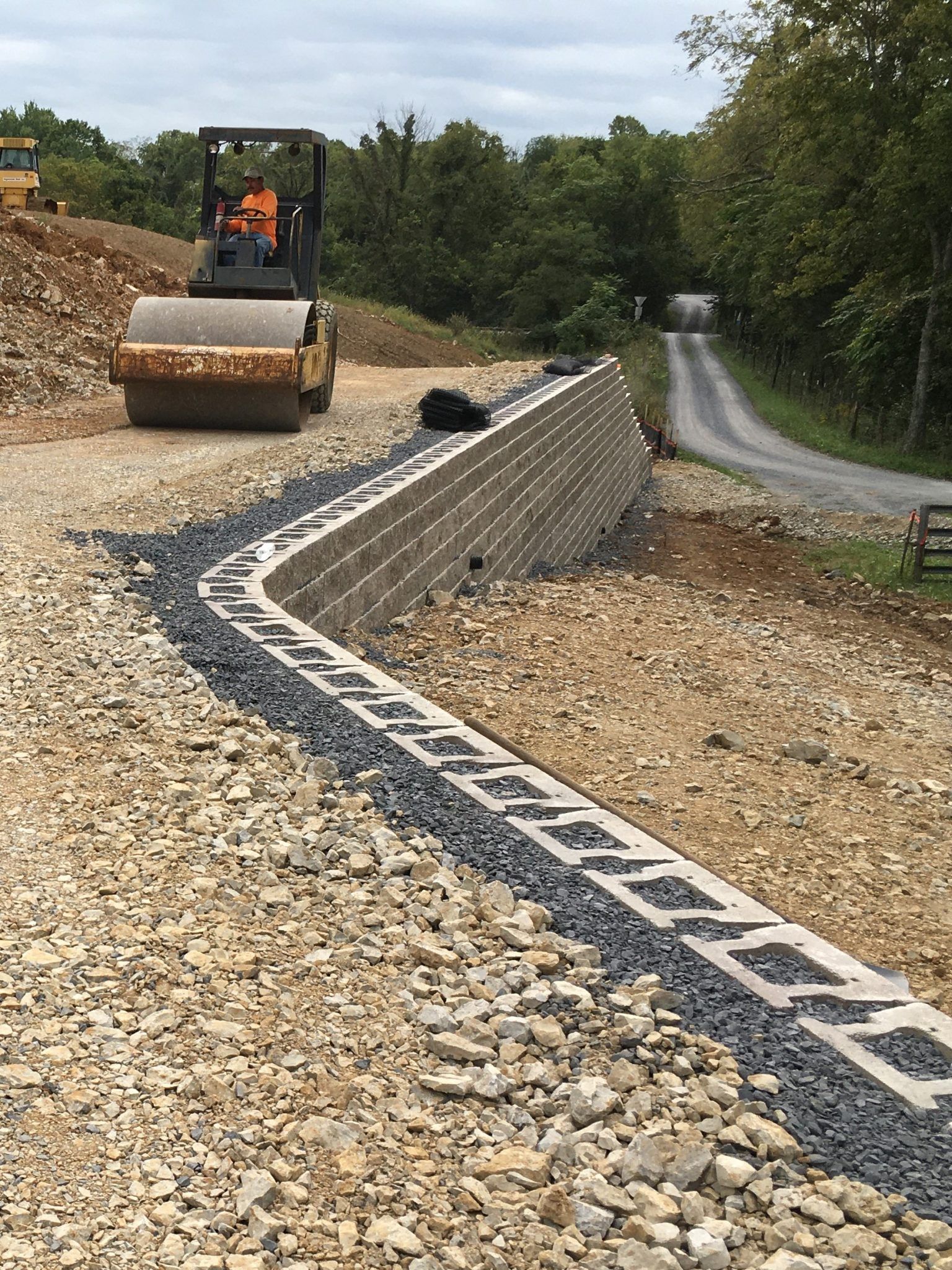 Construction site with a gravel road, retaining wall blocks, and a road roller in the distance
