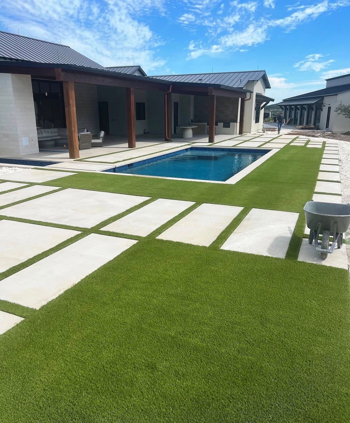 Modern backyard patio with a small blue pool, white stone pavers, and green lawn under a sunny sky