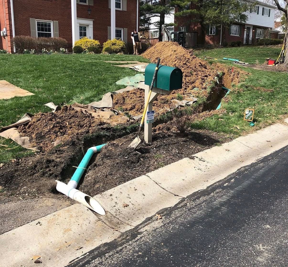 Broken green mailbox and exposed pipe beside a curb, with dirt and torn-up yard along a street.