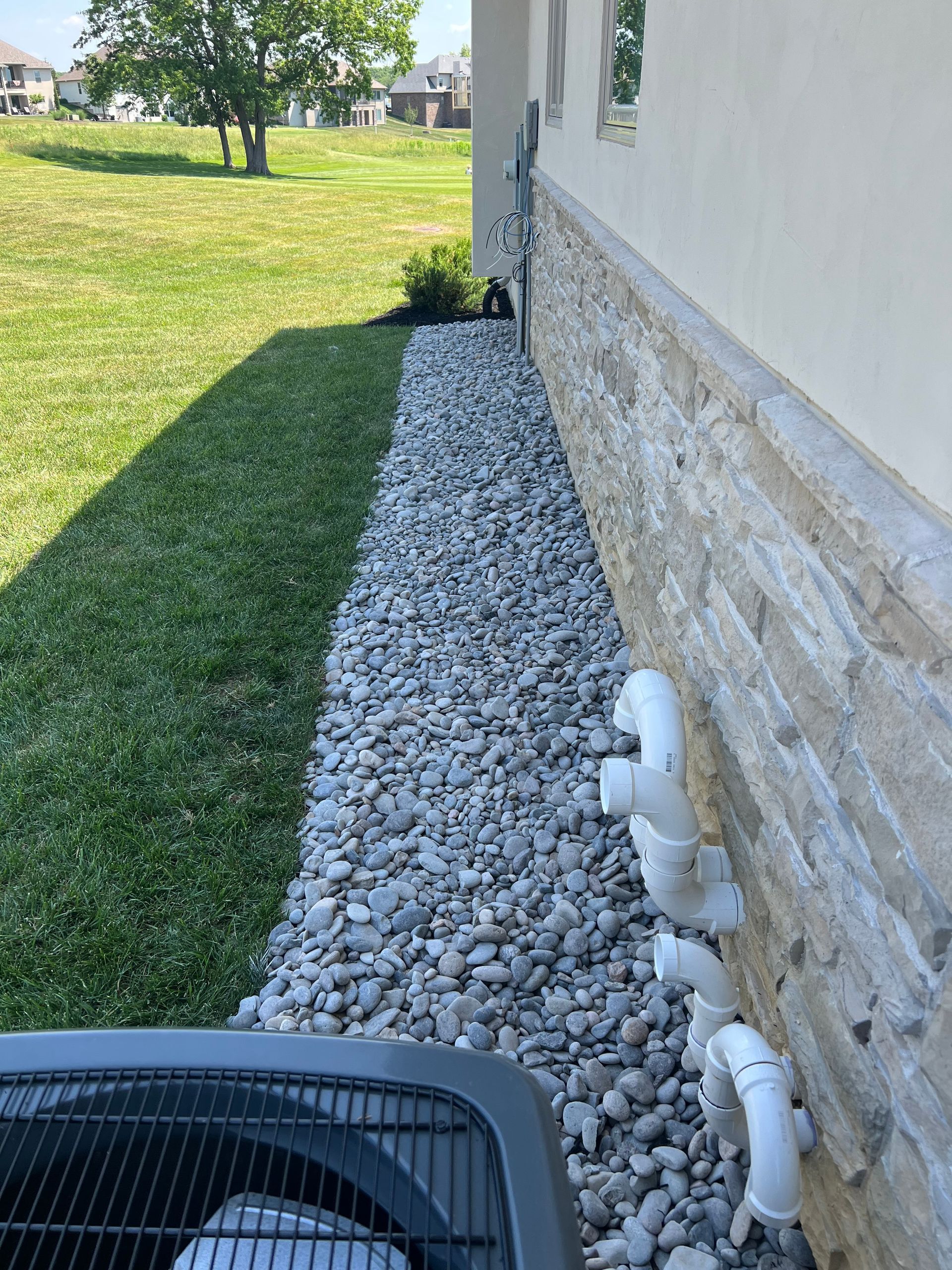 Gravel strip along house foundation beside green lawn, with white vent pipes and stone siding