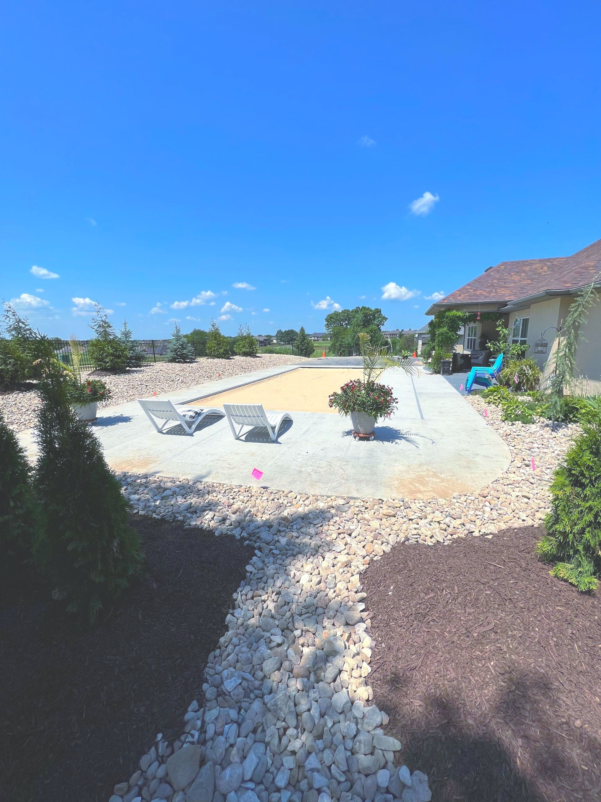 Backyard swimming pool with lounge chairs, potted plants, and a house under a bright blue sky