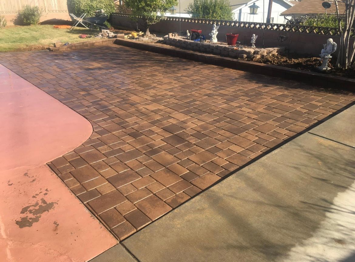 Brick-paved patio bordered by concrete and red concrete, with a landscaped edge and fence in the background