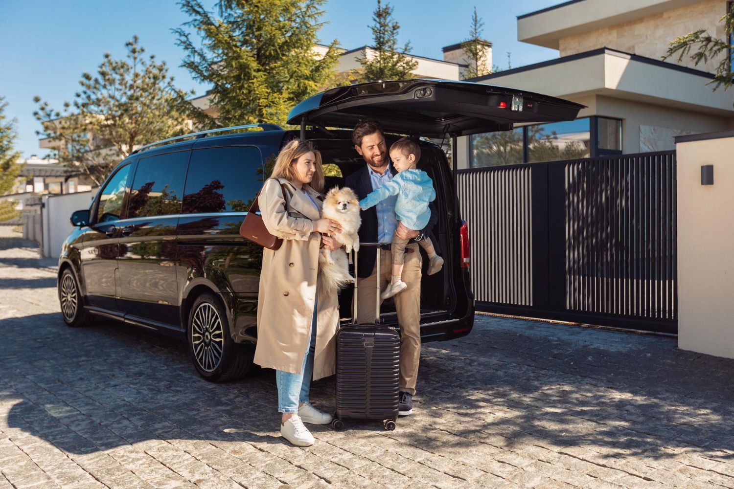 Family with luggage and dog loading into a black van in front of a house.