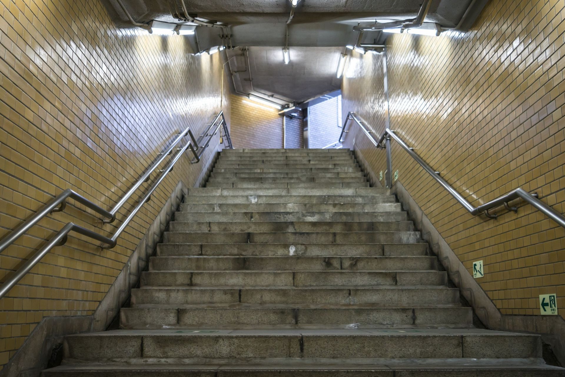 Staircase ascending into an underground tunnel, yellow tiled walls, metal handrails on either side.