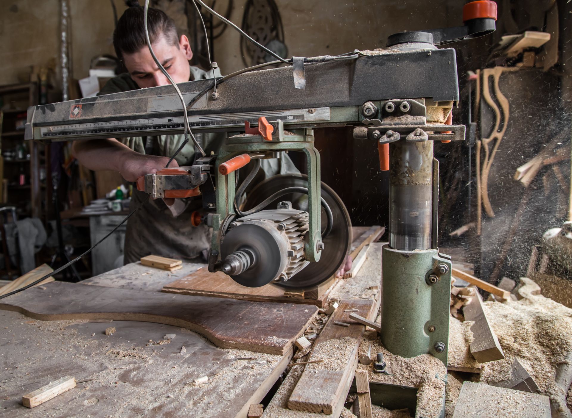 A man using a radial arm saw to cut wood in a workshop, sawdust flying.