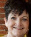 Woman with short, dark hair, smiling, wearing hoop earrings, brick background.