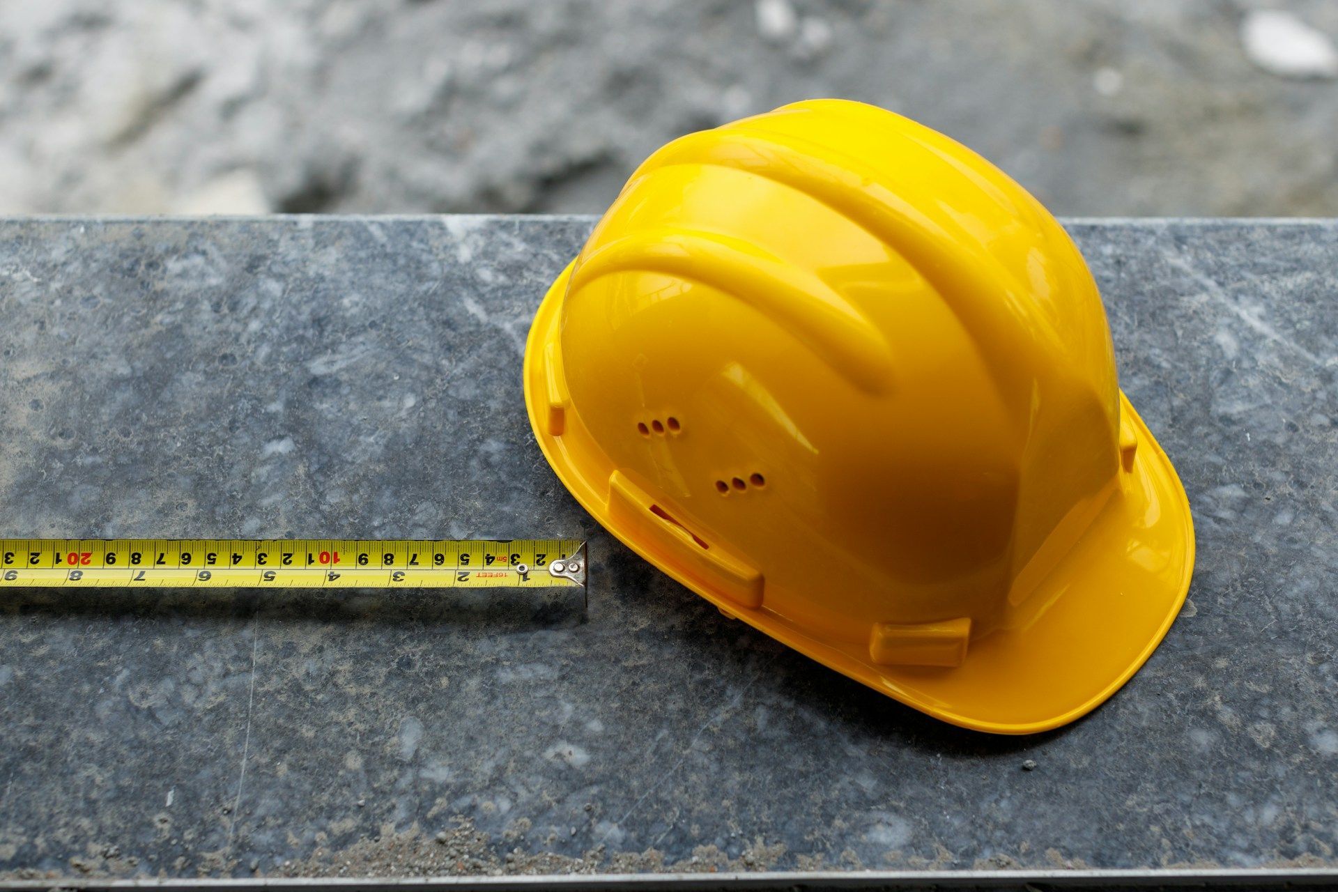 Yellow hard hat next to a measuring tape on a gray surface.