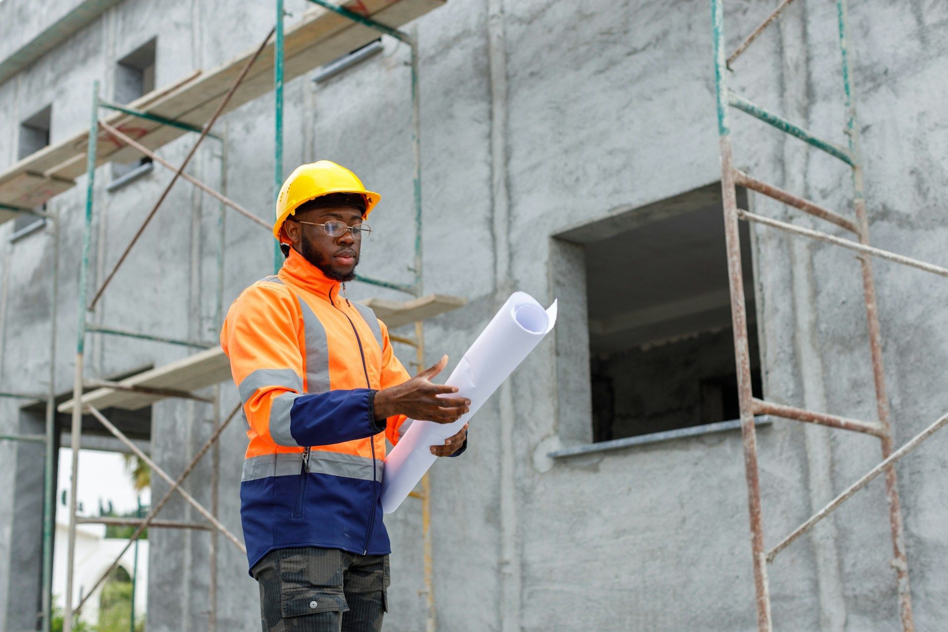 Construction worker in yellow hard hat and orange vest examining blueprints on a building site.