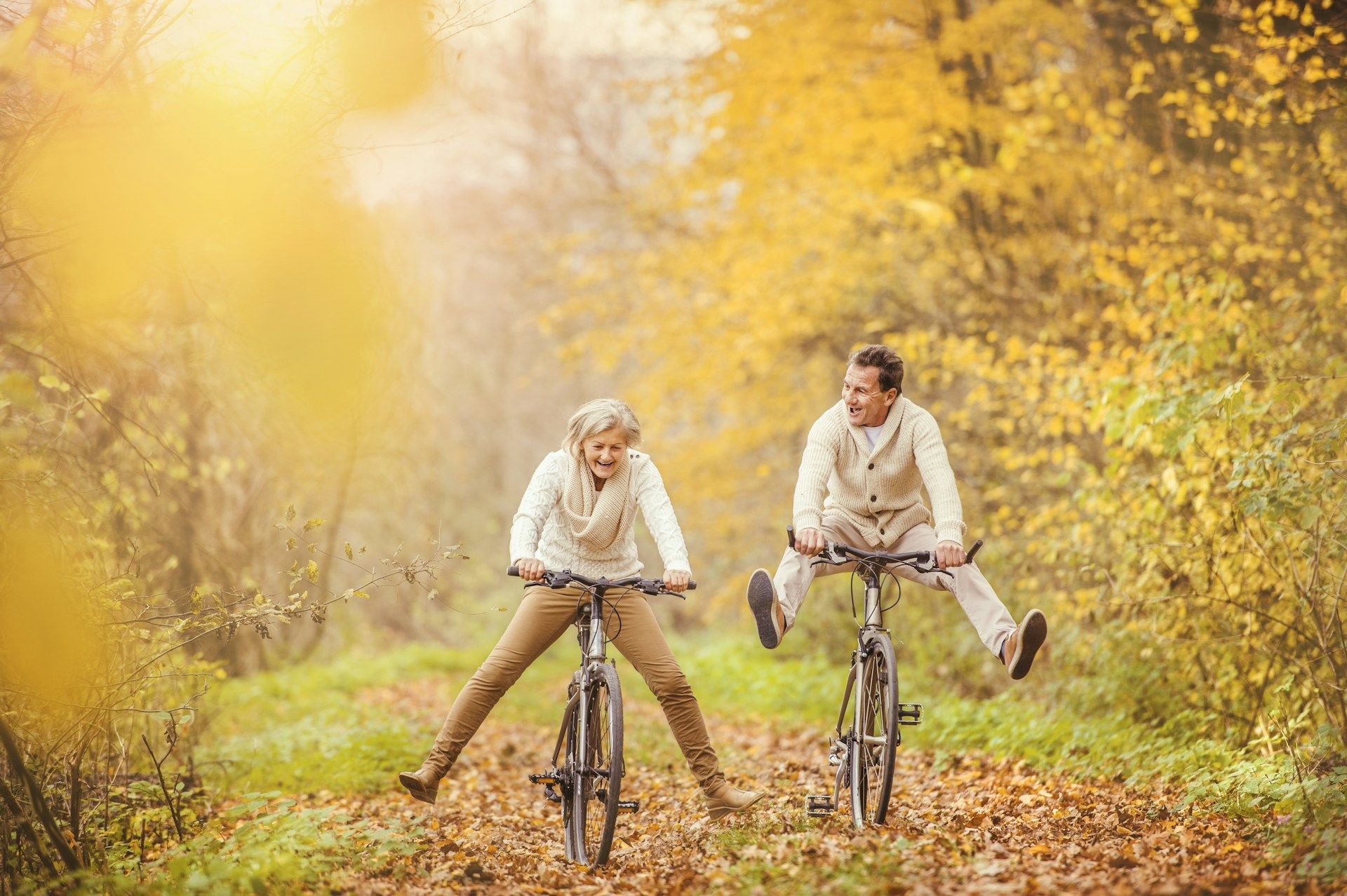 Couple joyfully riding bikes through a fall forest, legs spread wide. Golden foliage surrounds them.