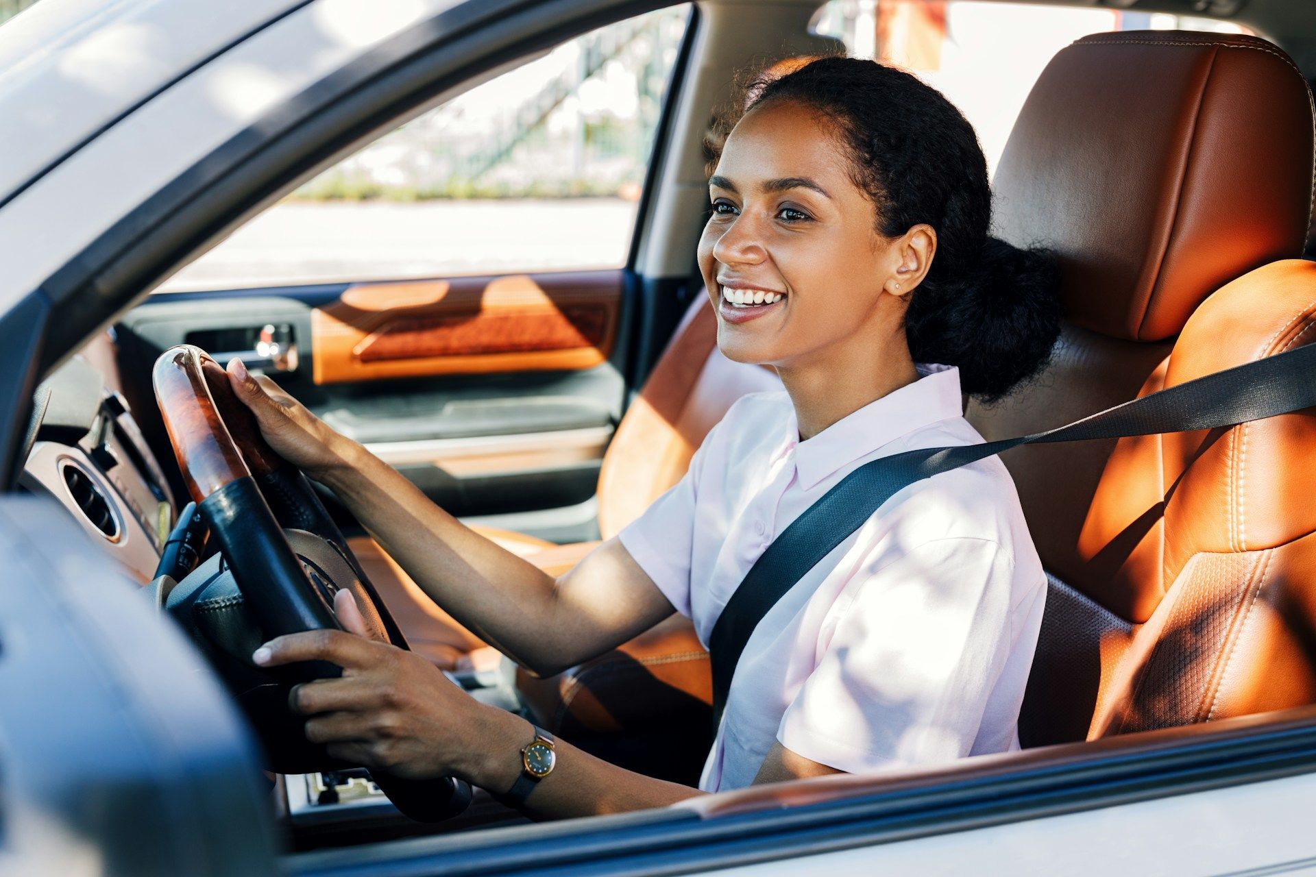 Woman smiling while driving a car, wearing a seatbelt, brown interior.