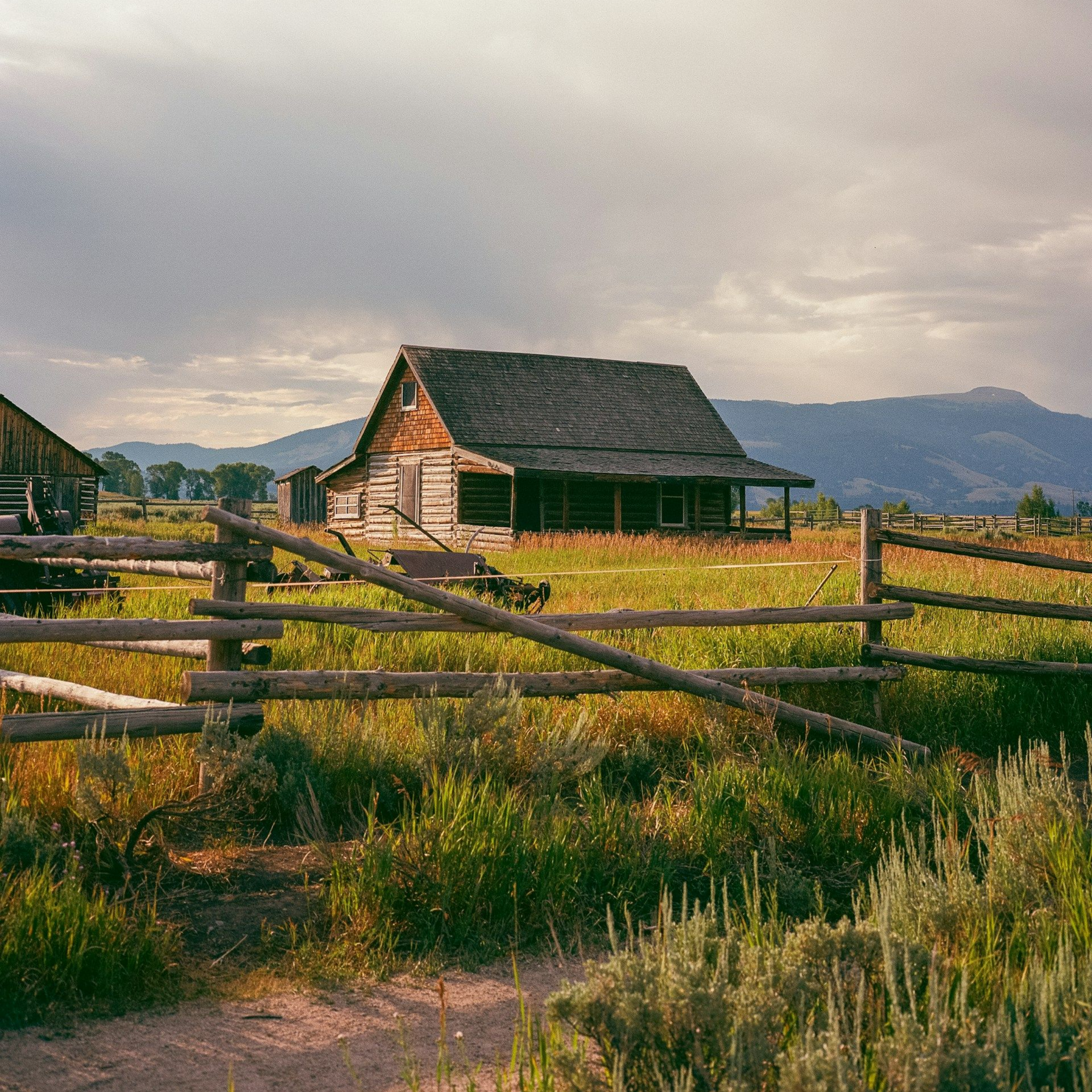 Rustic wooden cabin with a wooden fence and grassy field, mountains in the background. Cloudy sky.