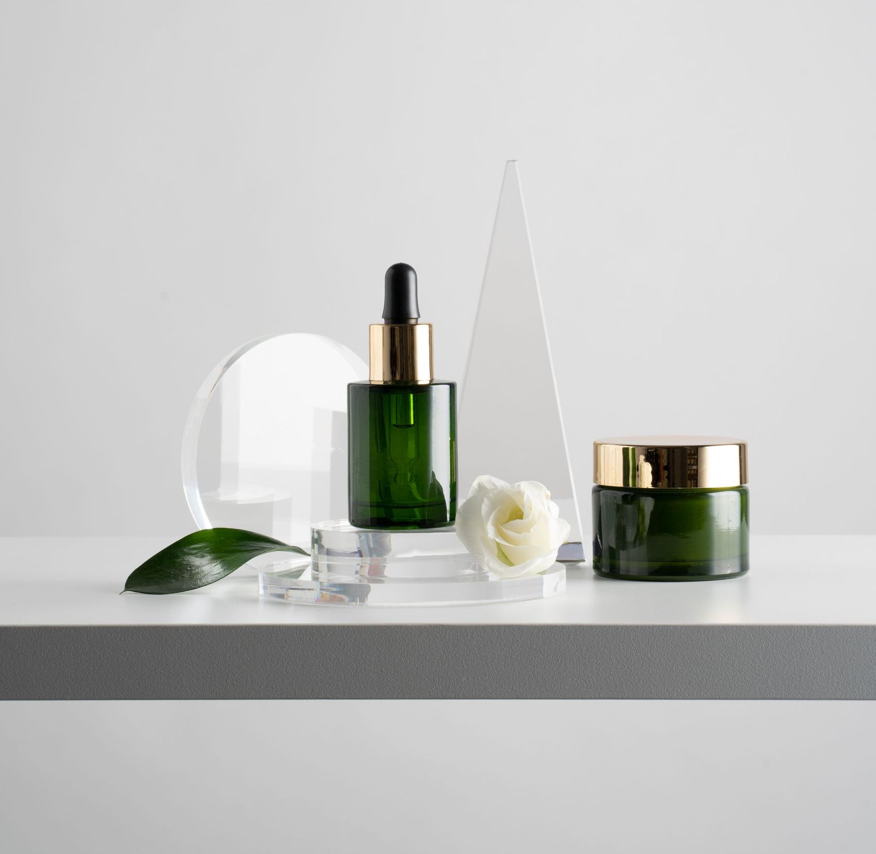 A bottle of serum , a jar of cream , and a rose are on a table.