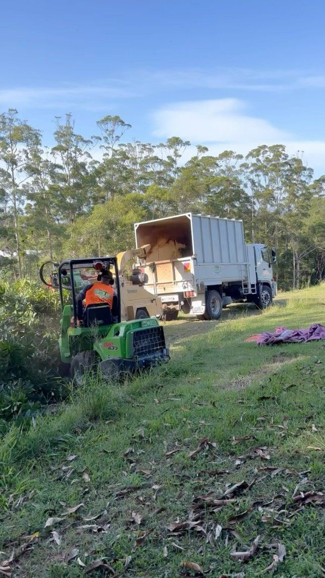 A Man Is Driving a Tractor Next to A Truck in A Field — Hinterland Tree & Stump Removal in Palmview, QLD