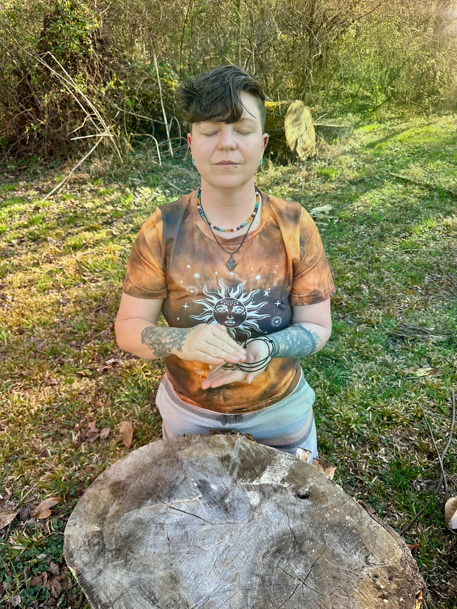 Woman sitting cross-legged in meditation, outdoors. Wearing linen clothes, jewelry, eyes closed, hands clasped.
