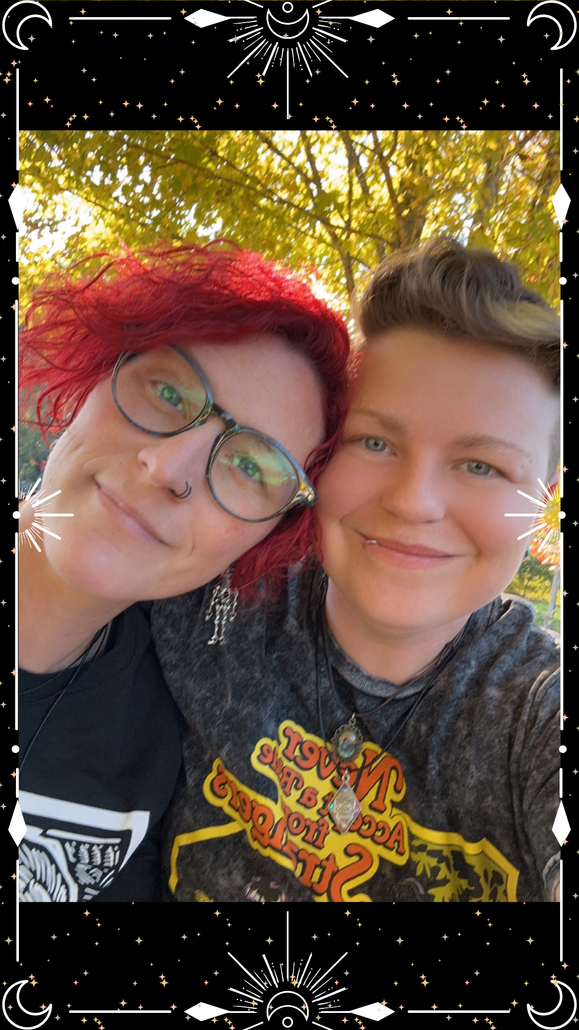 Two people smile close together, one with red hair, glasses, the other with short hair, both with light skin.