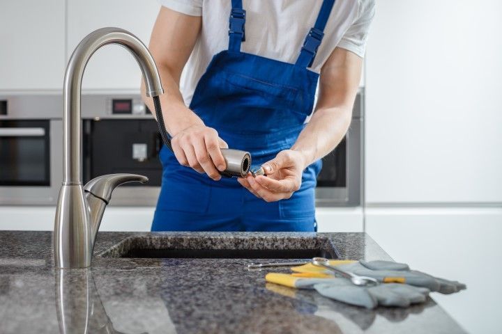 Person in blue apron washing dishes at a kitchen sink with soap and sponges on the counter.