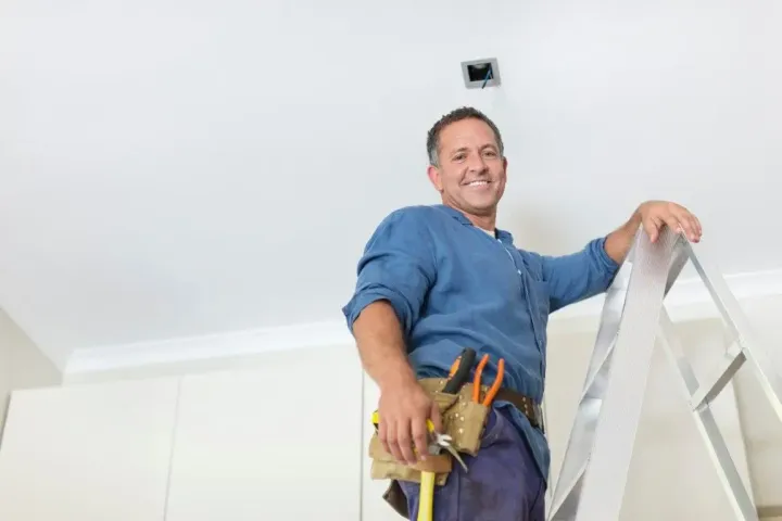 Smiling worker on a ladder, holding tools and painting supplies in a bright room