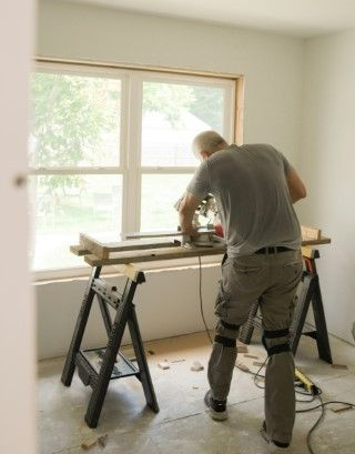 Person cutting a wood board on sawhorses in a bright room with a large window