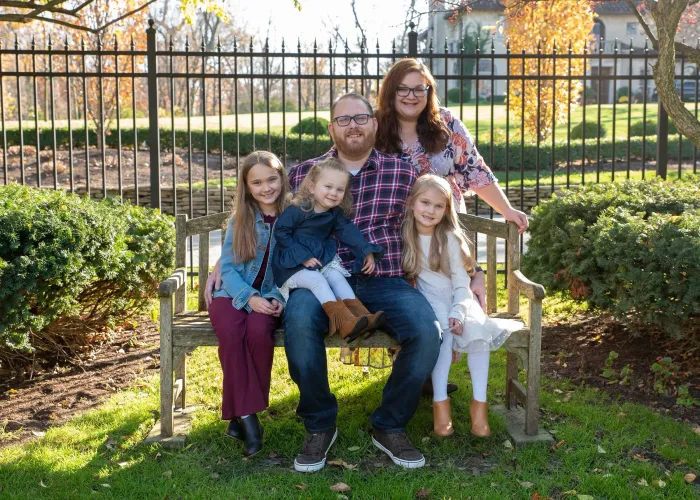 Family seated on a bench outdoors in a sunny park, smiling at the camera.