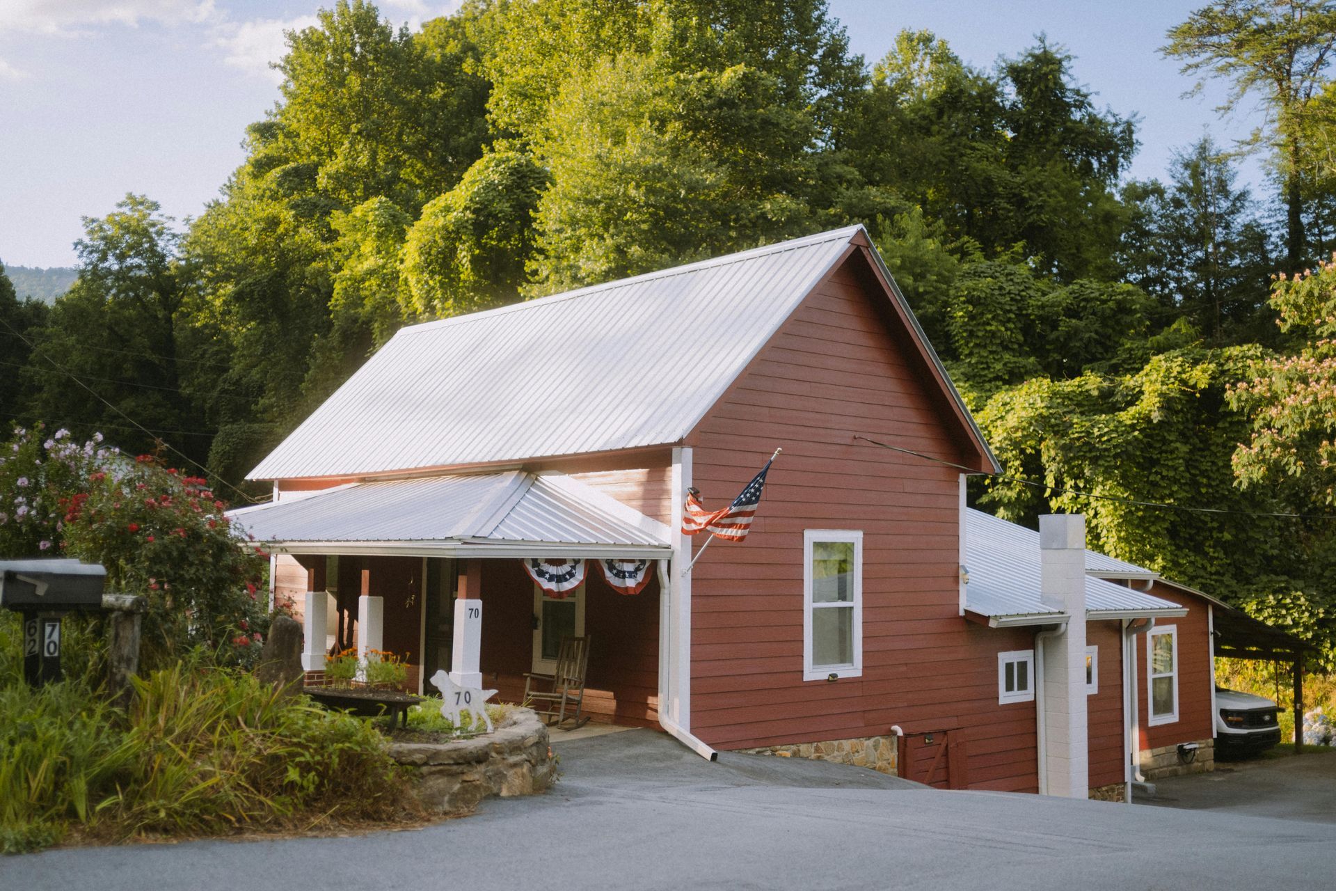 Red brick house with white porch and metal roof, surrounded by trees and greenery