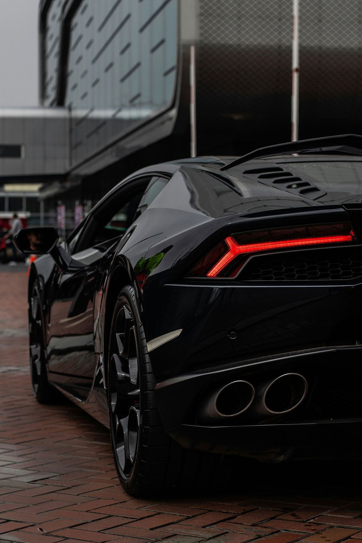 Black Lamborghini parked on brick; red taillight illuminated, modern building background.