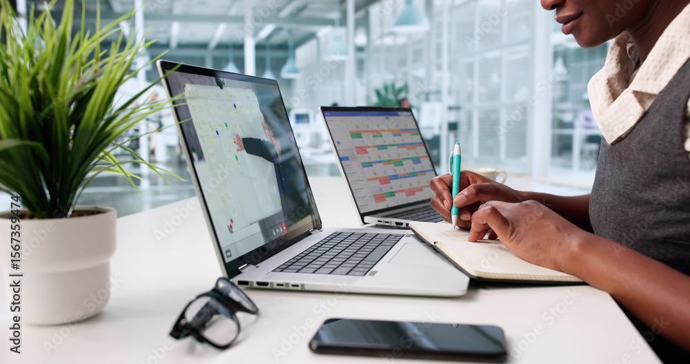 Woman taking notes while watching a presentation on a laptop, with a second laptop showing graphs, indoors.