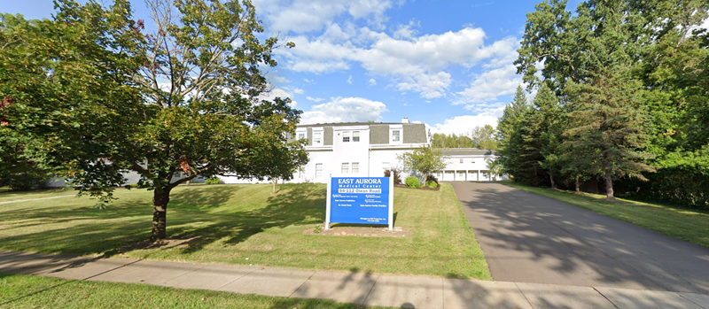 A white building with a blue sign in front, trees on either side, and a driveway.