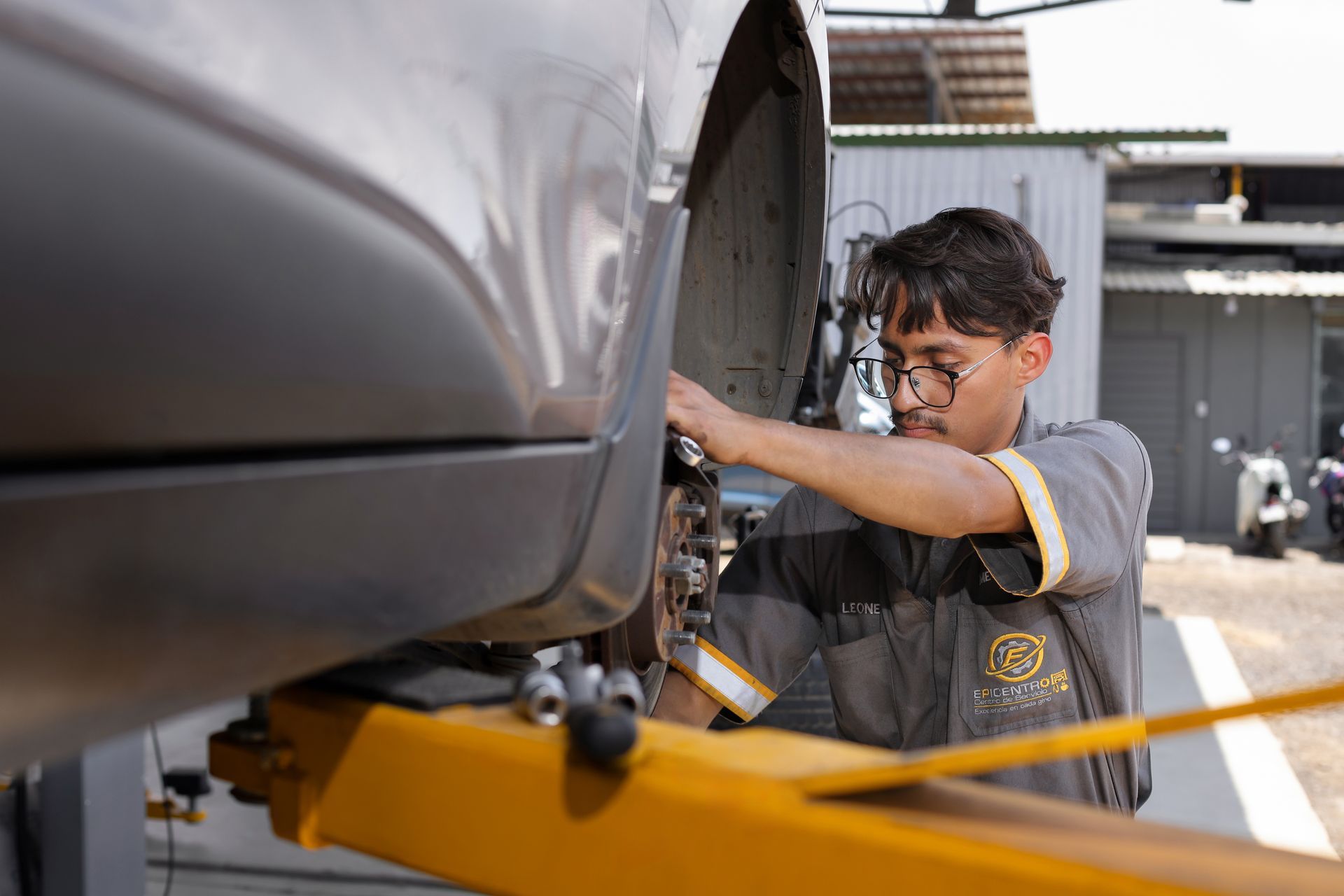 Mecánico inspeccionando la rueda y el sistema de frenos expuestos de un coche plateado en un taller de reparación.