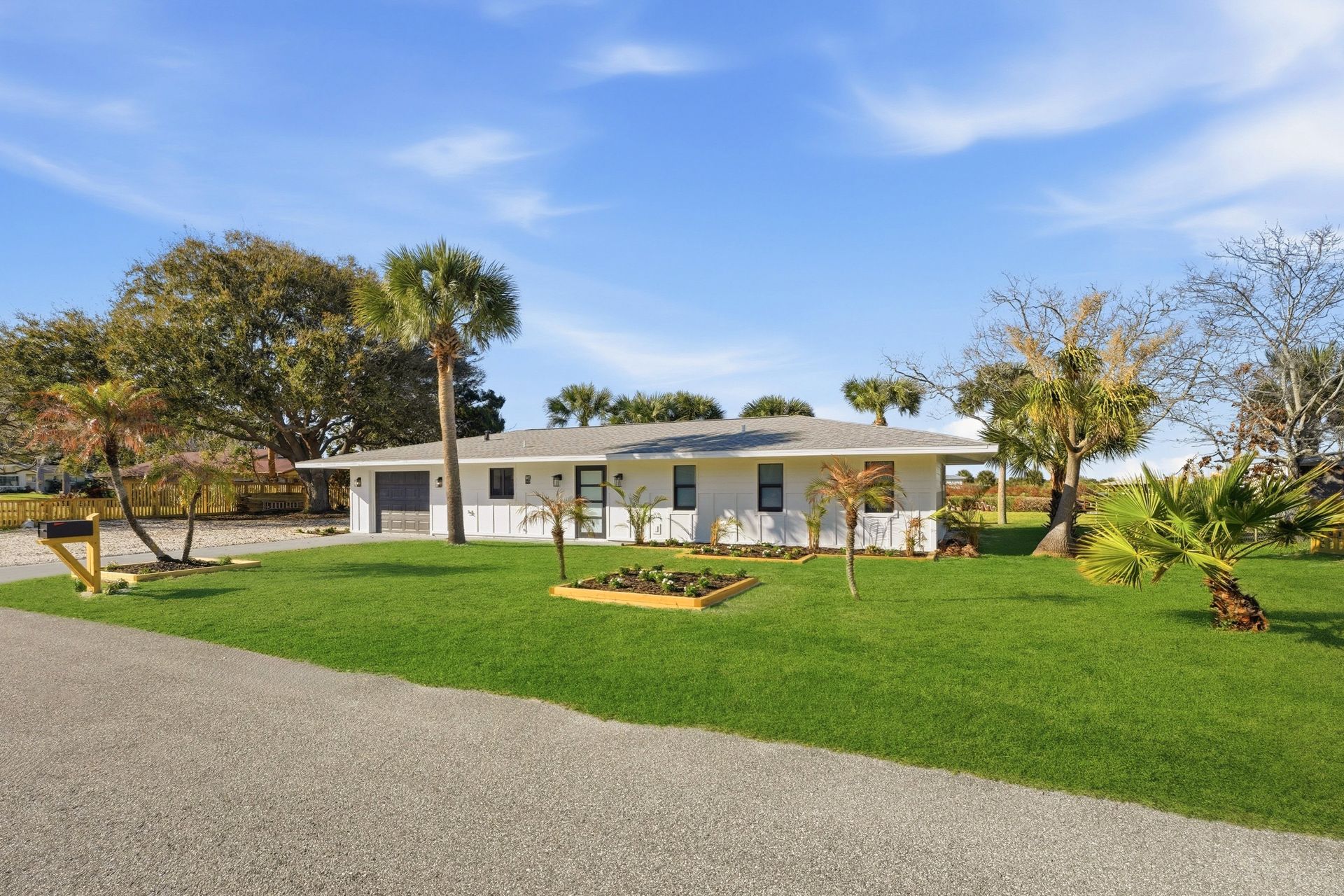 A single-story white house with a grey roof sits on a green lawn with trees under a bright blue sky.