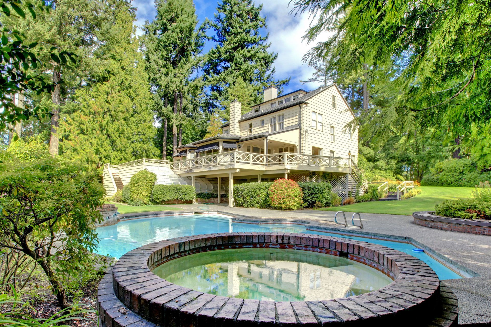 A large, light-colored house with a deck overlooking a pool and a circular brick hot tub surrounded by lush trees.