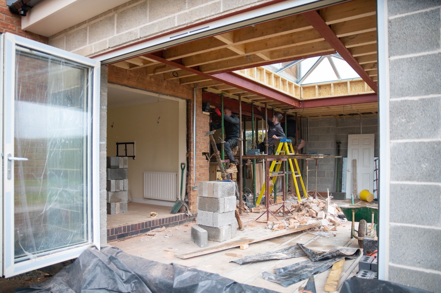 Workers renovating a residential extension, with exposed beams and scaffolding visible in the open construction site.
