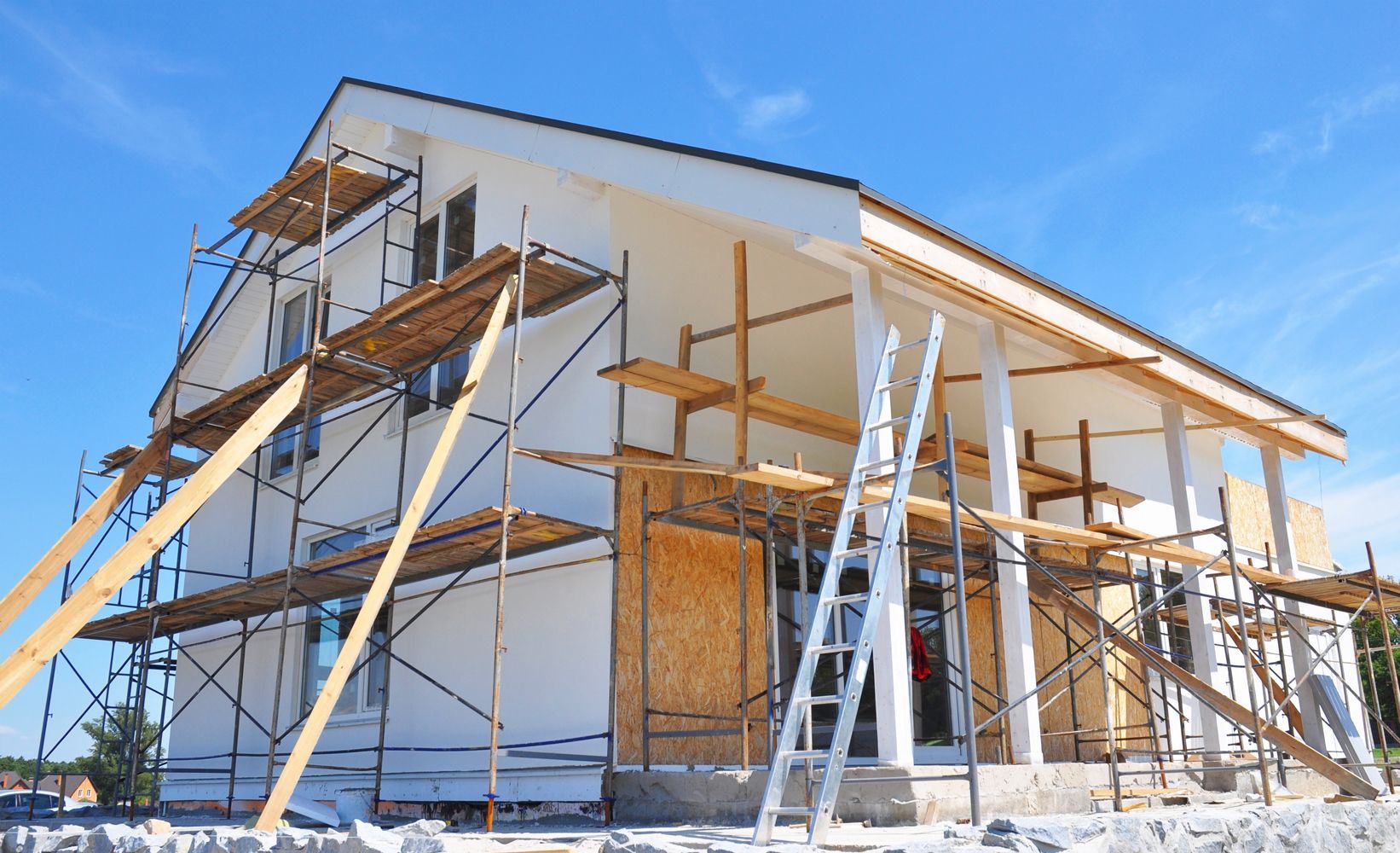 A house under construction with external scaffolding, white walls, and wood paneling under a clear blue sky.