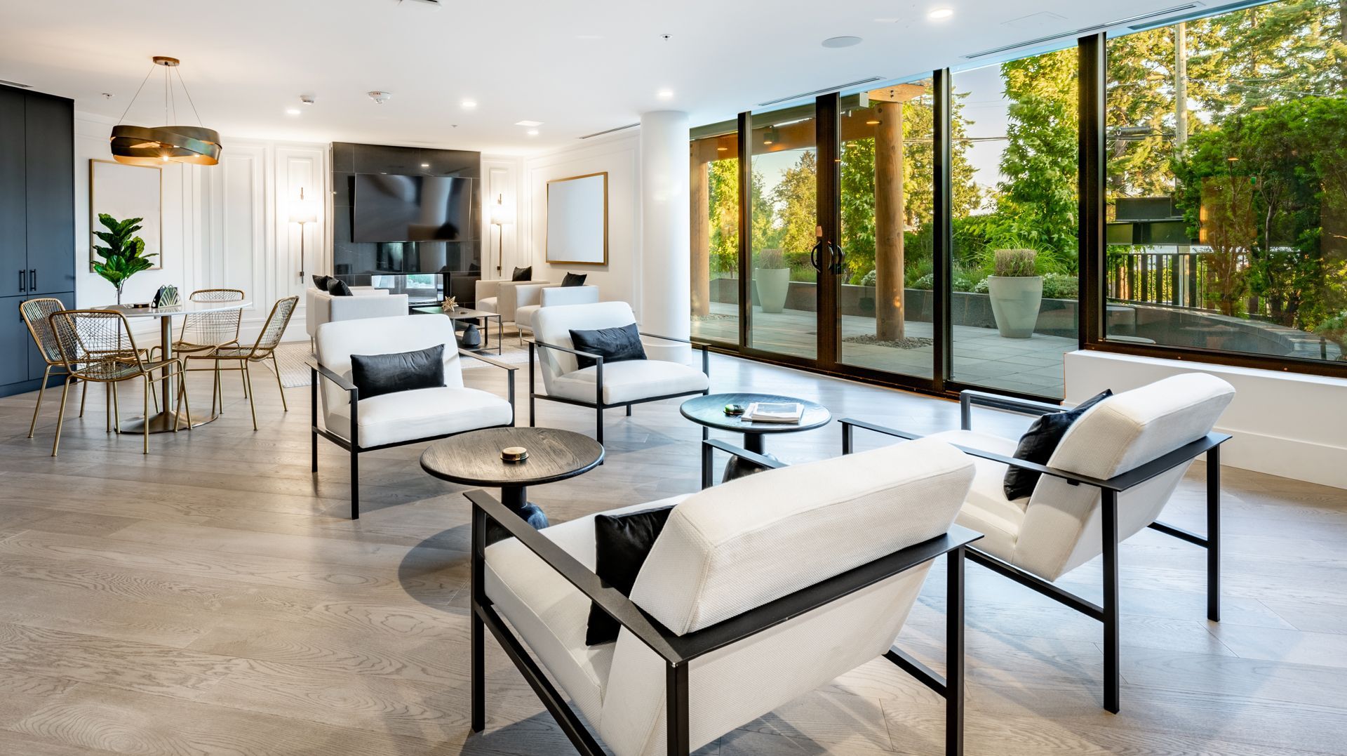 A modern, sunlit lounge featuring white armchairs with black accents, small circular tables, and large glass windows.