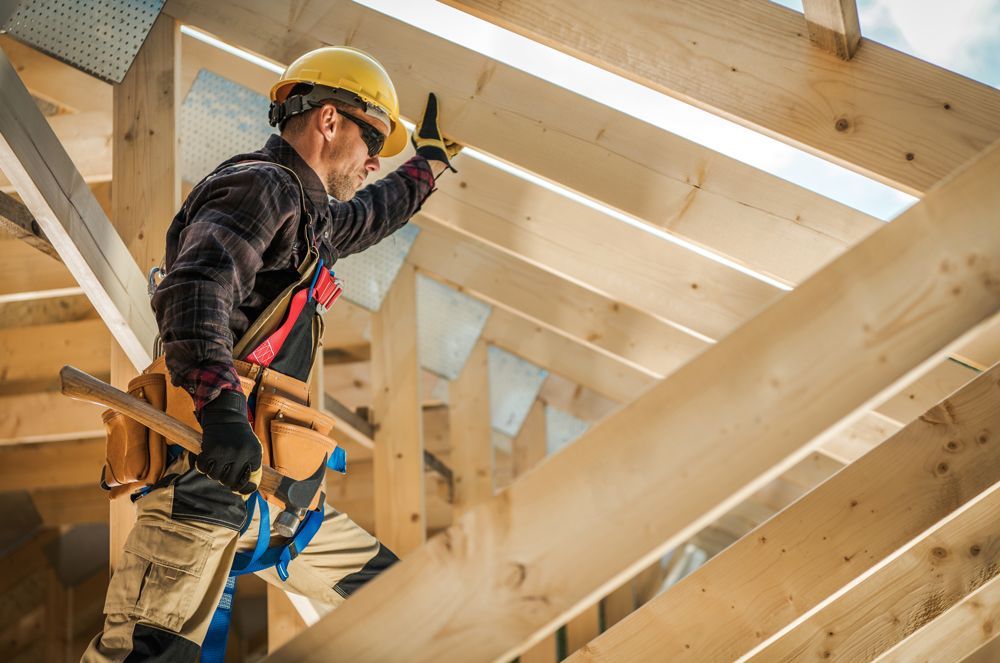 A worker in a hard hat and tool belt stands on a wooden frame, inspecting exposed rafters at a construction site.